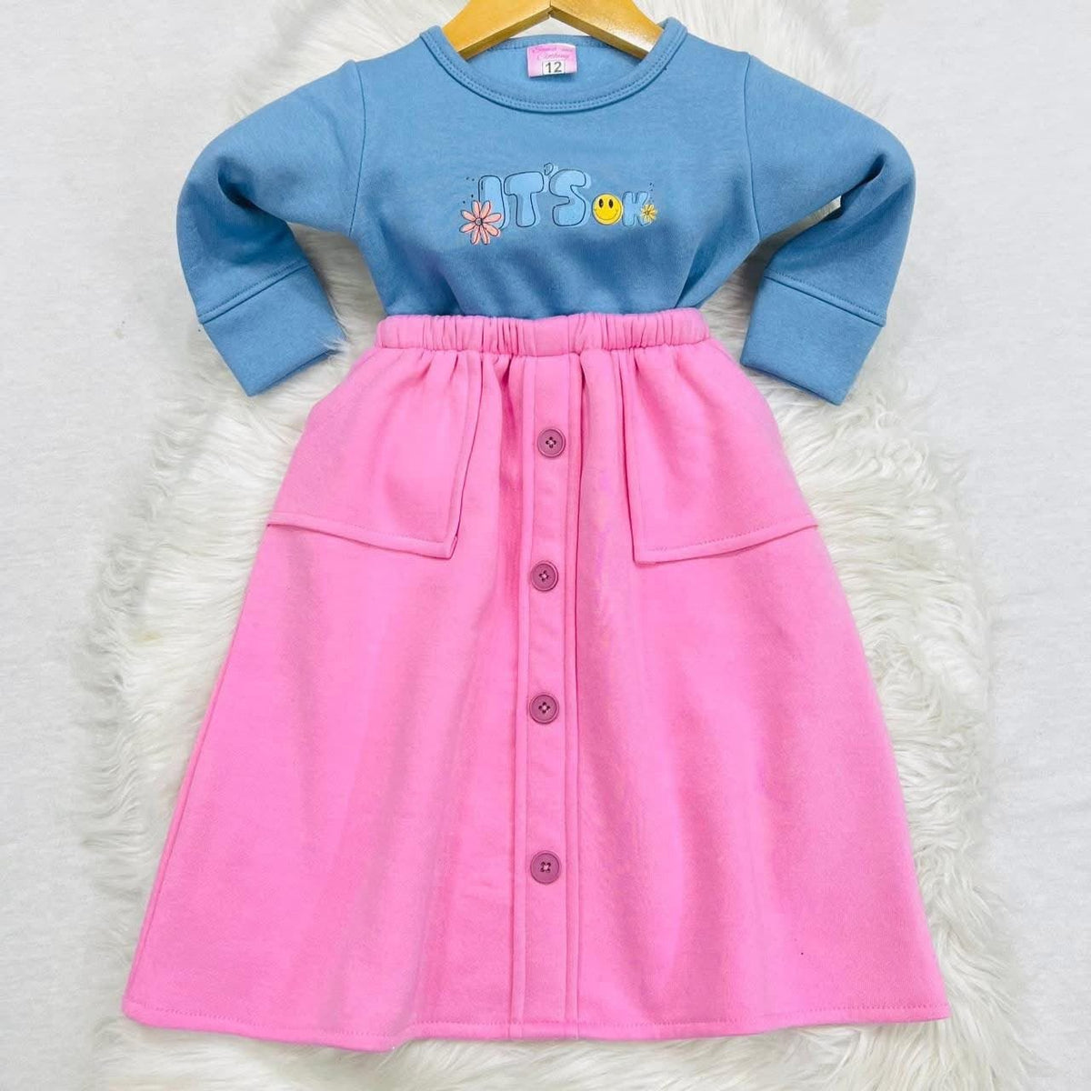 A light blue long-sleeve top with an "IT'S OK" graphic paired with a pink button-front midi skirt